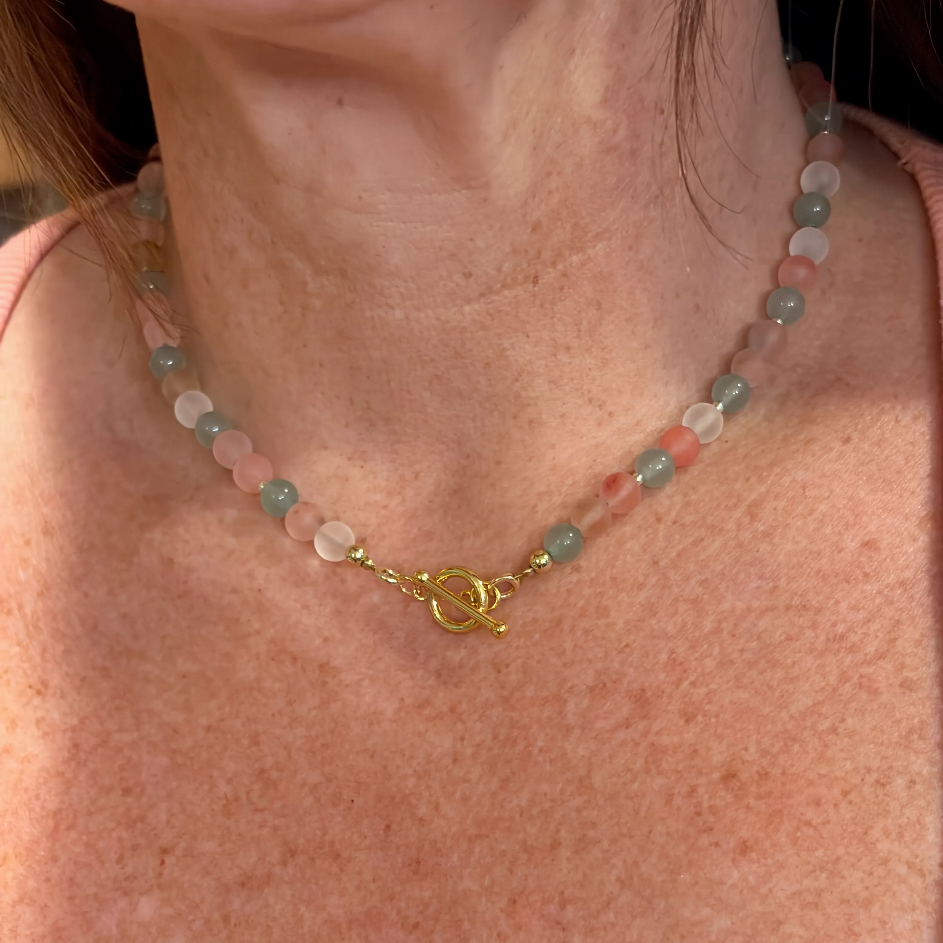 Woman wearing a beaded necklace with frosted pink and green gemstones and a gold toggle clasp.