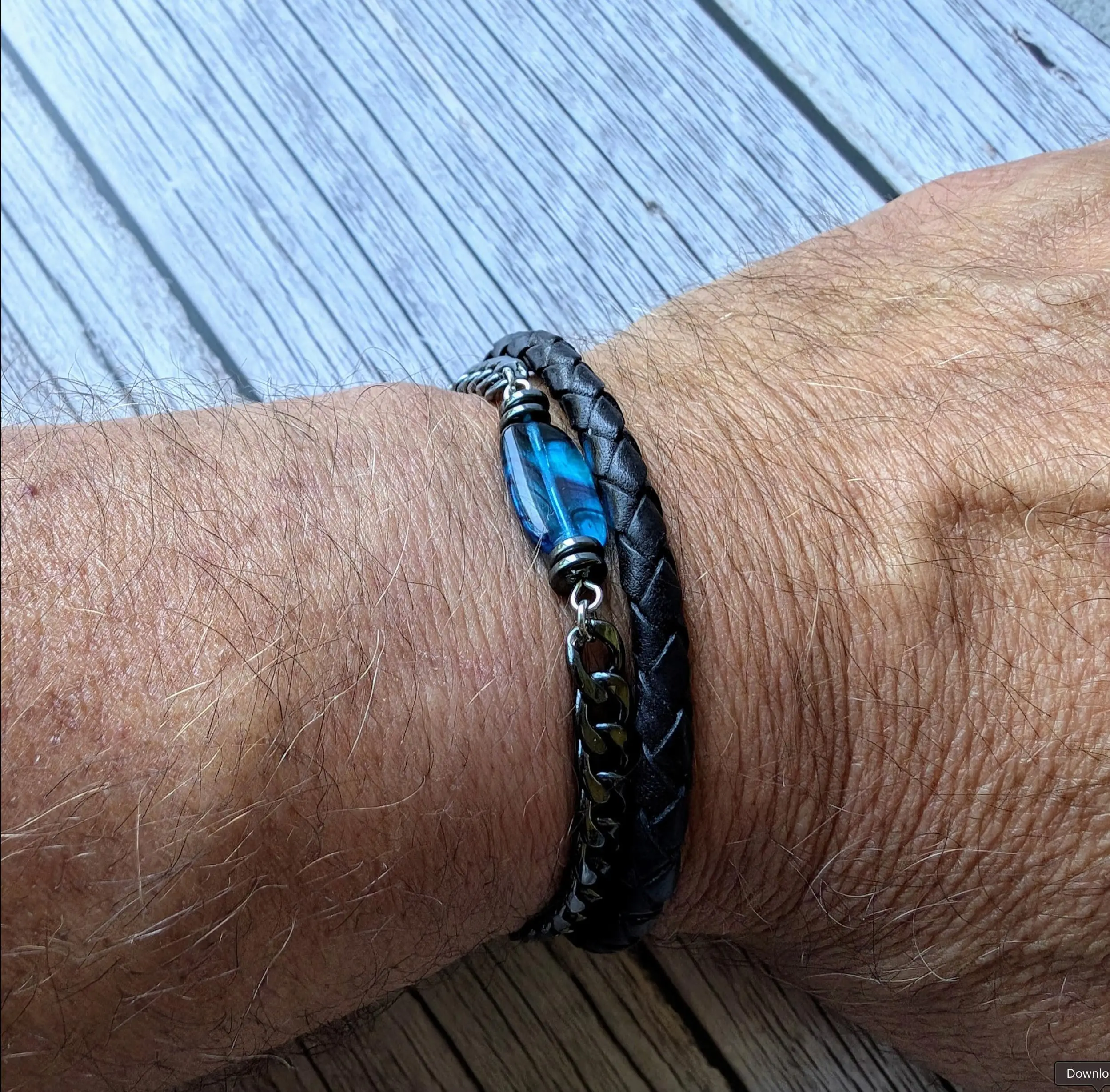 Black leather braided bracelet with blue striped cylindrical bead and metal accents worn on a man's wrist.