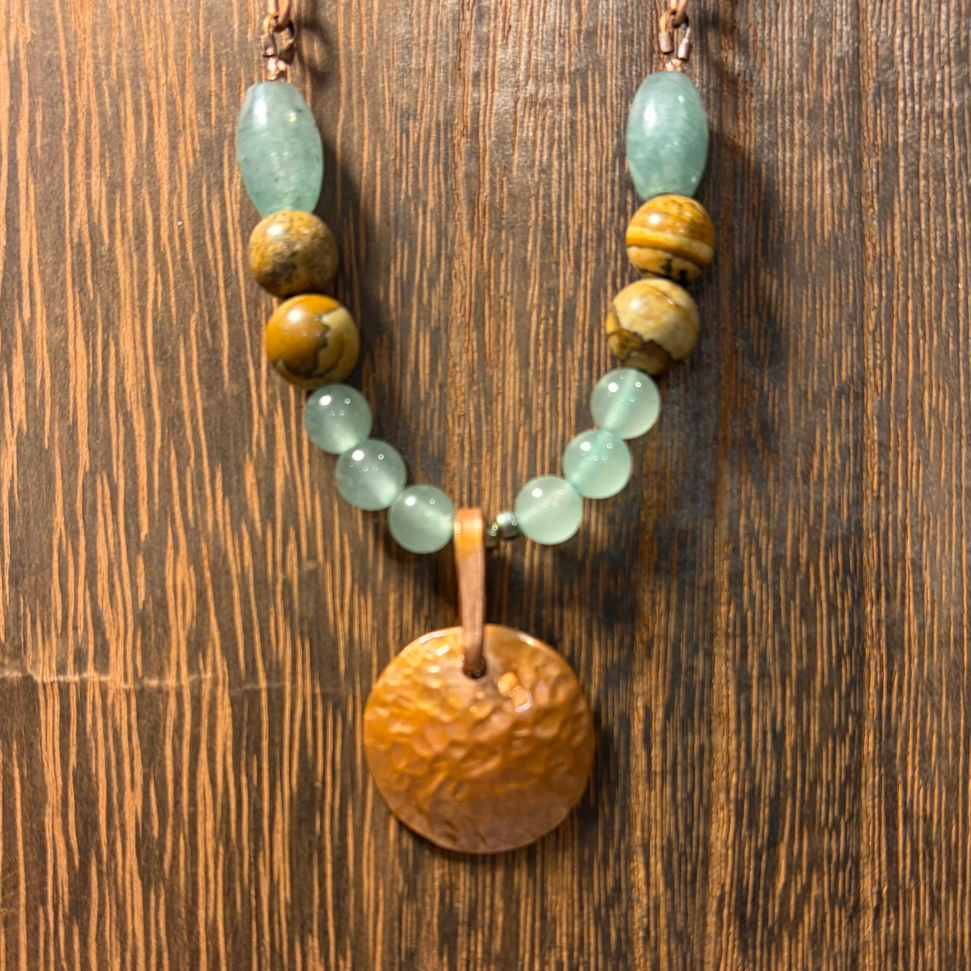 Handmade necklace with pale green jade beads, tiger's eye stones, and a textured copper pendant on a leather cord.