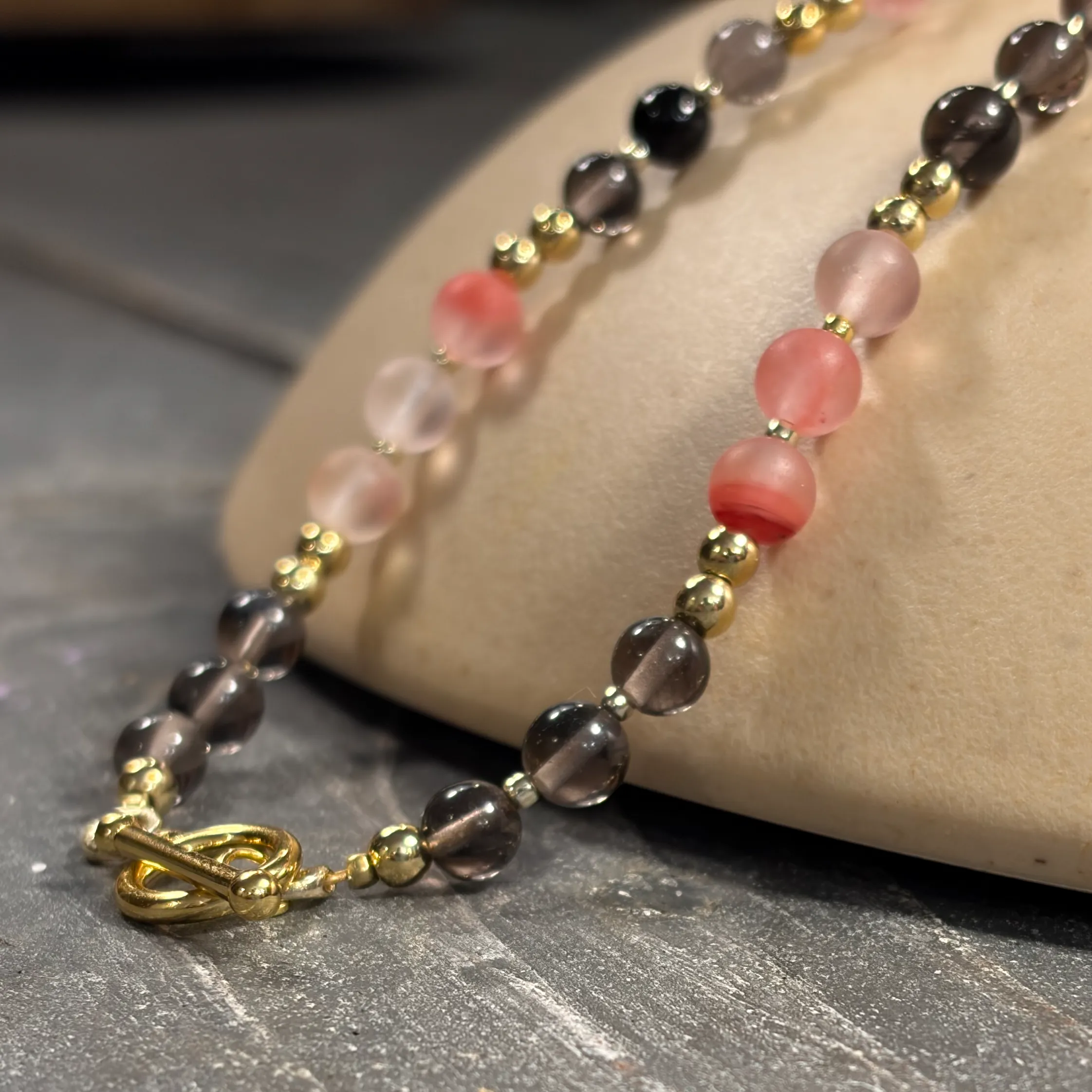 Two beaded bracelets with rose quartz and smoky quartz stones separated by gold-tone spacer beads, displayed on a cream-colored stand.