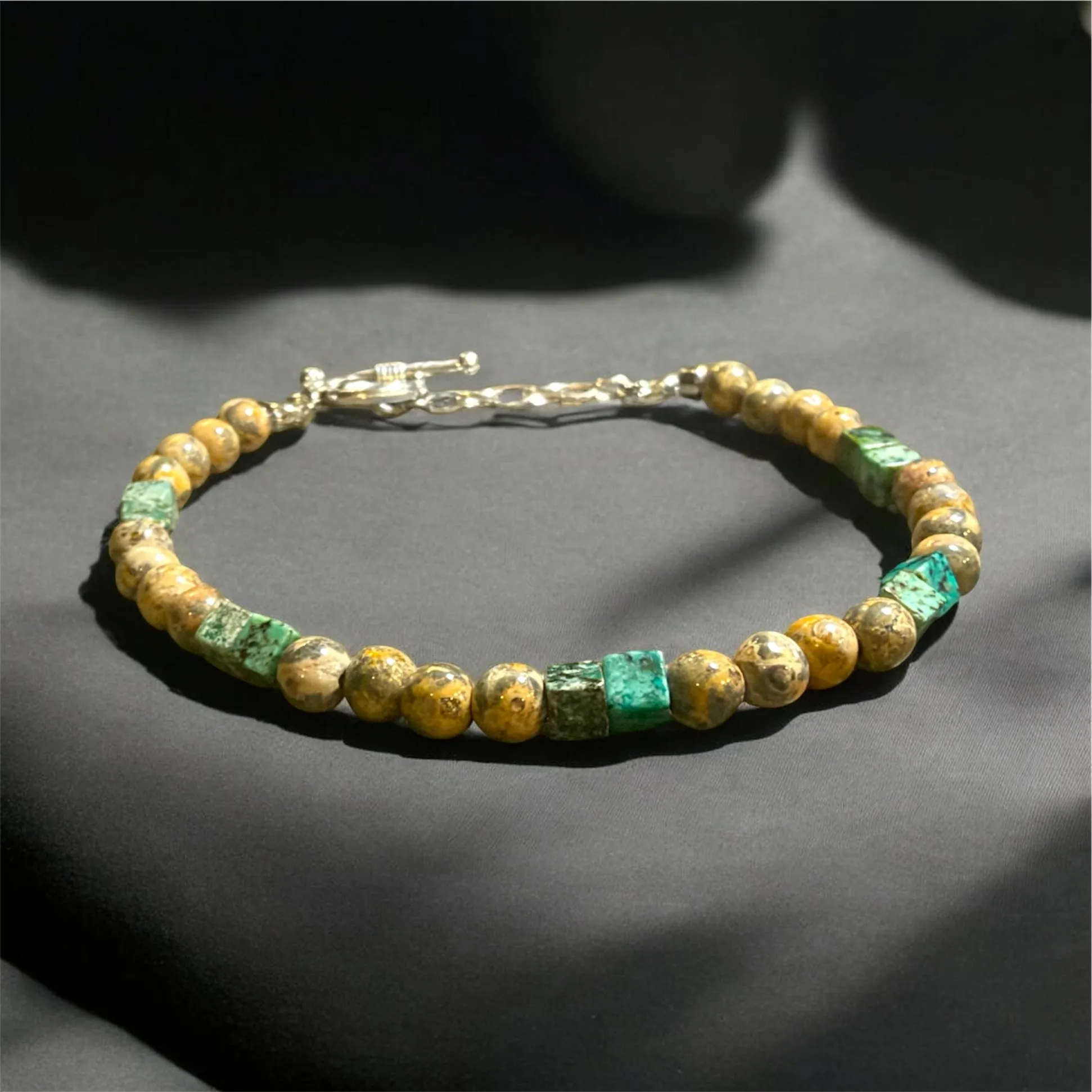 Gold-toned beaded bracelet with green emerald cube accents and gold toggle clasp on gray surface.