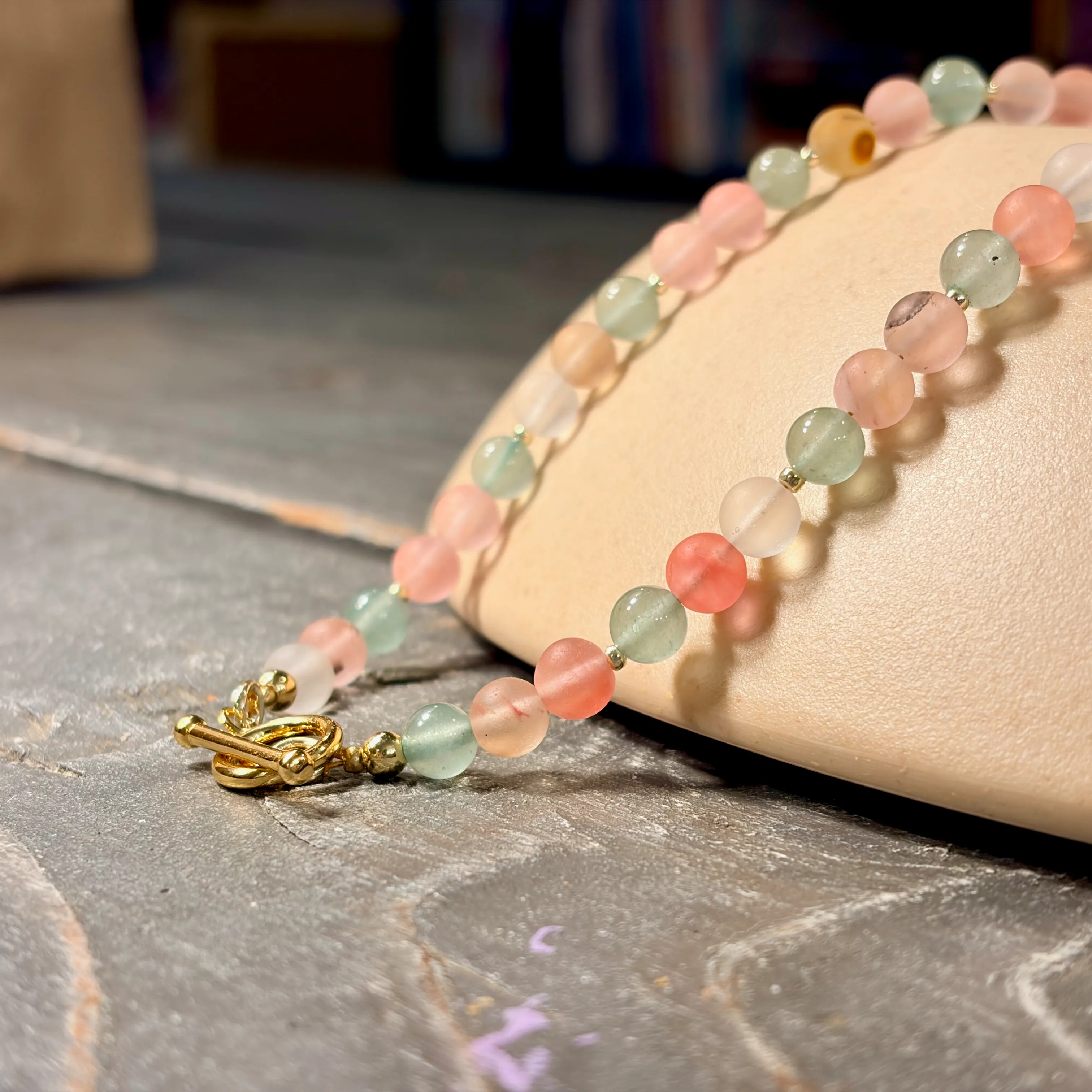 Beaded bracelet with pale pink, mint green, and cream-colored stone beads with gold-tone clasp displayed on a cream ceramic dish.