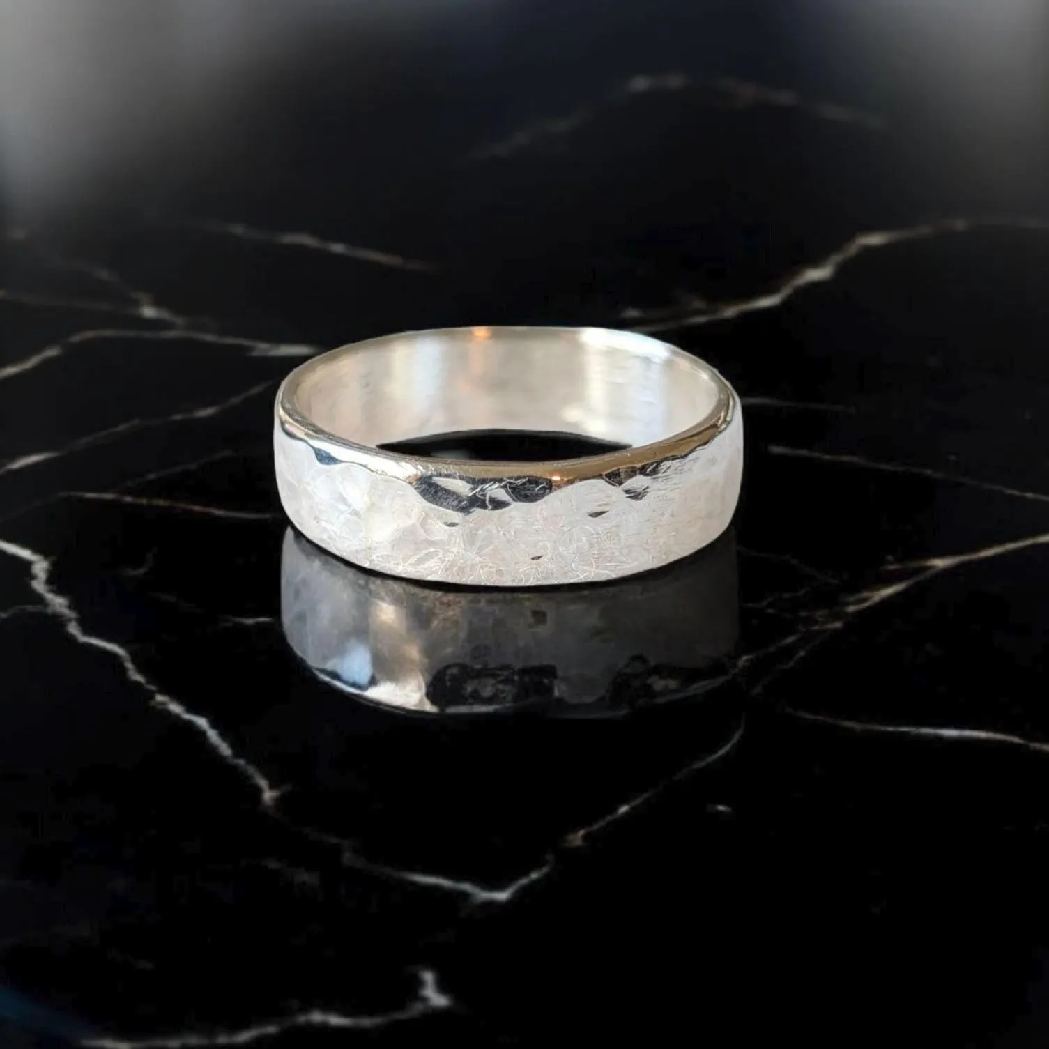Silver band ring with textured hammered finish displayed on black marble surface.