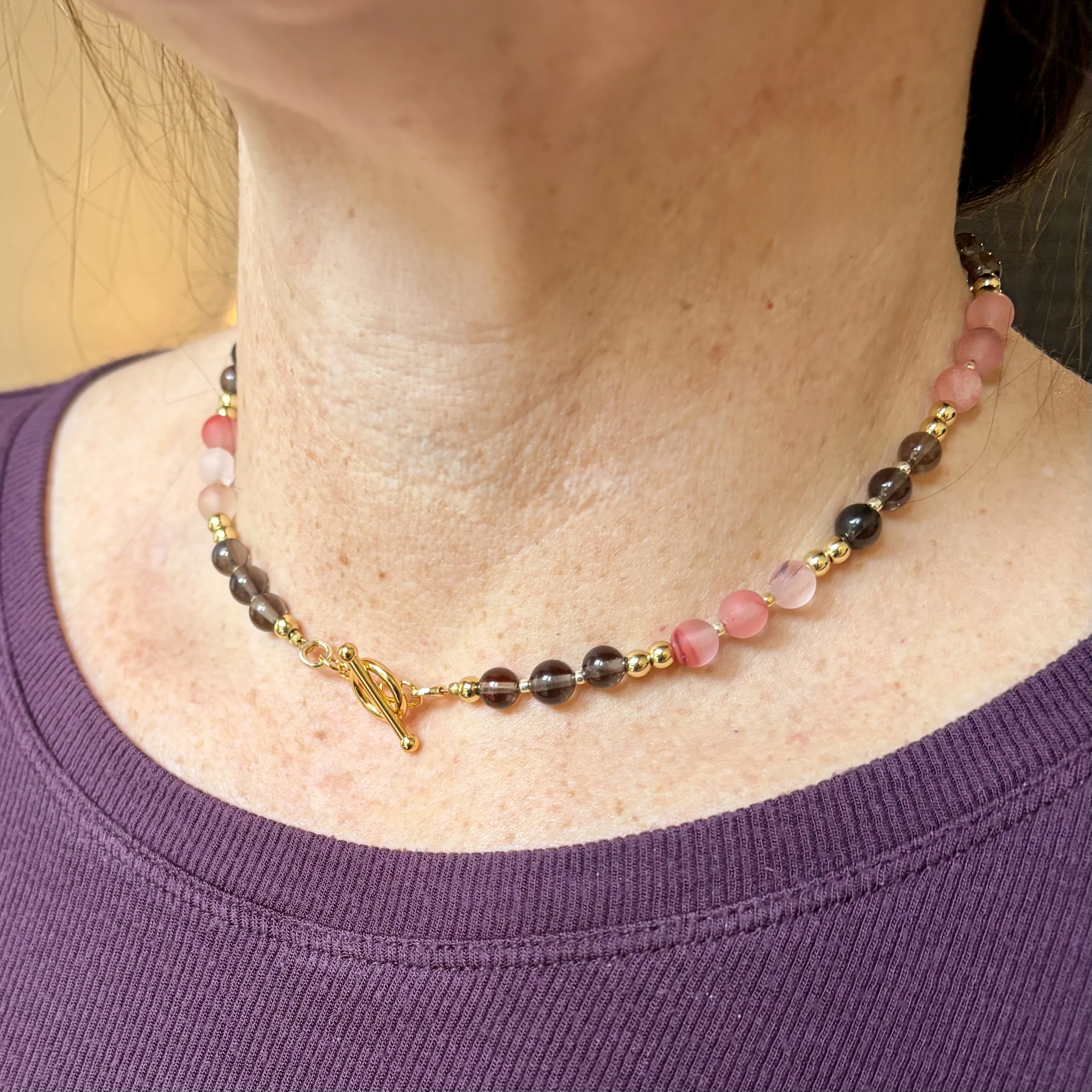 Woman wearing a beaded necklace with smoky quartz and pink tourmaline stones separated by gold spacer beads and a gold toggle clasp.