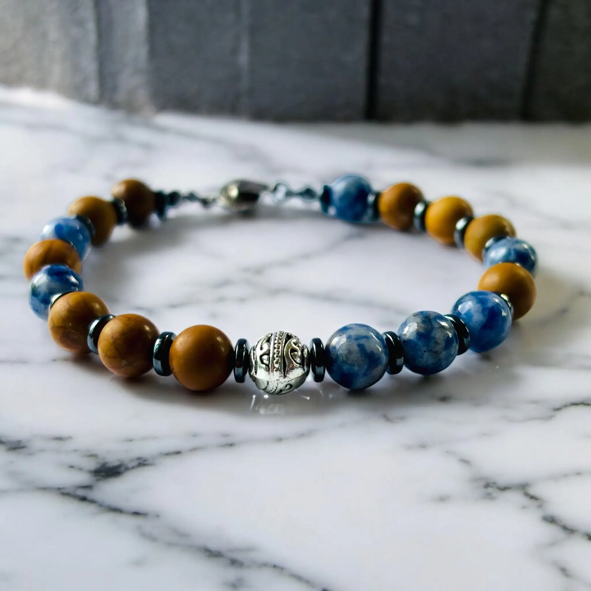 Beaded bracelet with alternating golden and blue stone beads separated by black spacers and a decorative silver centerpiece on marble background.