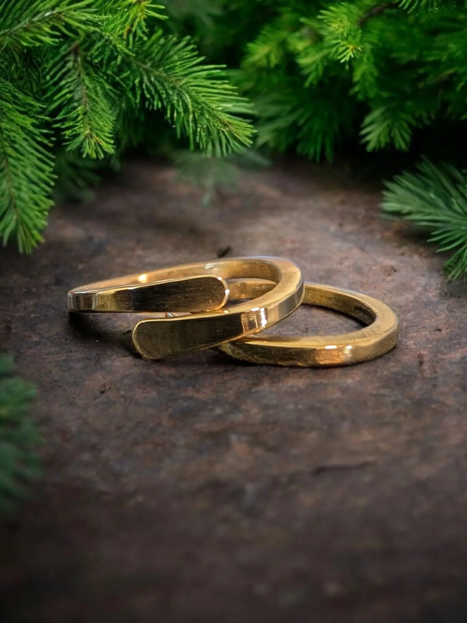 Two polished gold wedding bands displayed on a dark stone surface with evergreen branches above.
